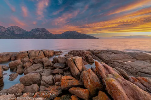 Lichen-streaked granite boulders in warm light at Coles Bay, with apricot and violet clouds over calm water and the Hazards in the distance.