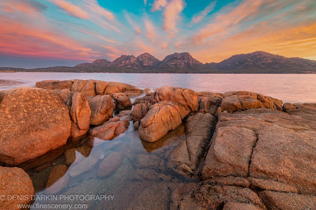 A tidal pool between granite boulders reflects a pink and blue sunset sky at Coles Bay, the Hazards peaks rising in the distance.