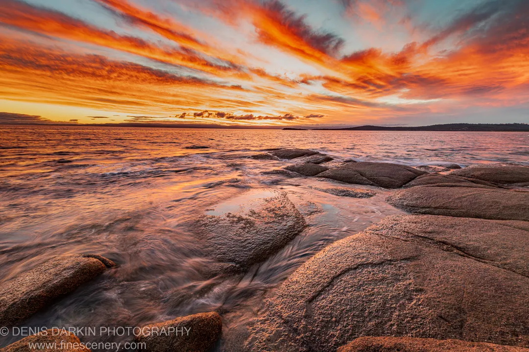 Wet granite boulders catch the warm glow of the setting sun at Coles Bay as gentle surf reflects in the wide-angle foreground.