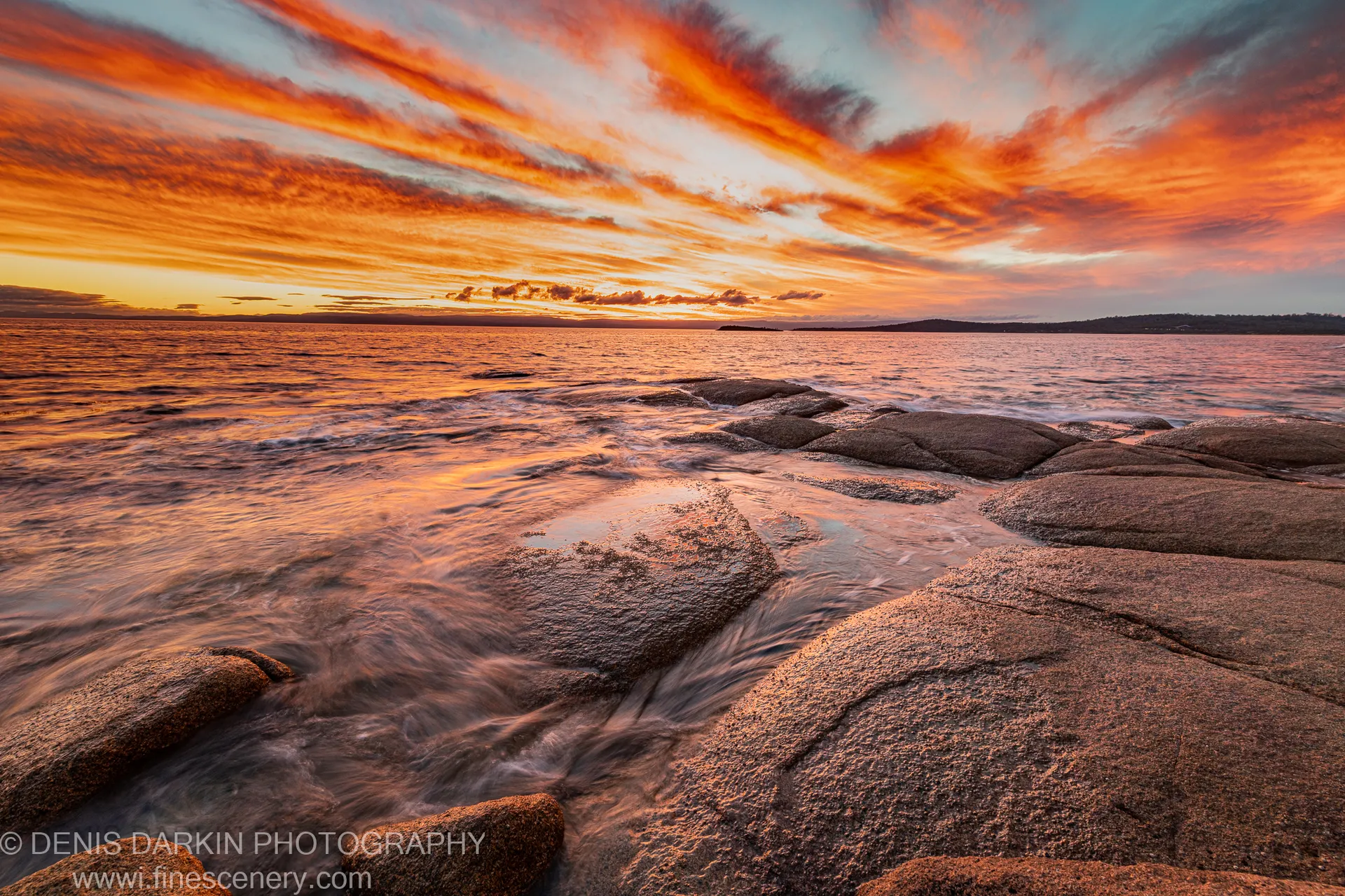 Wet granite boulders catch the warm glow of the setting sun at Coles Bay as gentle surf reflects in the wide-angle foreground.