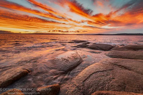 Wet granite boulders catch the warm glow of the setting sun at Coles Bay as gentle surf reflects in the wide-angle foreground.