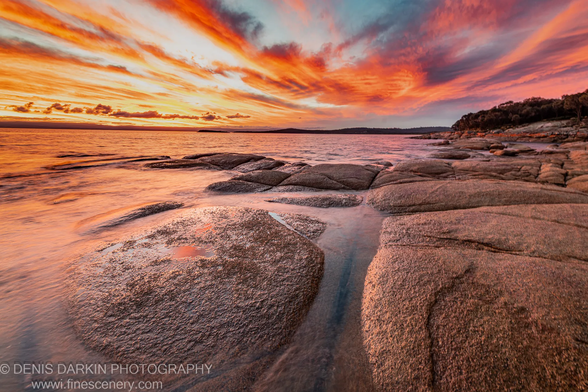 Long-exposure ocean water weaves between barnacle-covered granite boulders on a Tasmanian coastal shore as orange and pink clouds streak across the sunset sky.