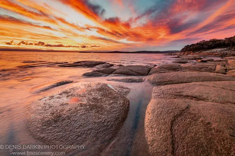 Long-exposure ocean water weaves between barnacle-covered granite boulders on a Tasmanian coastal shore as orange and pink clouds streak across the sunset sky.