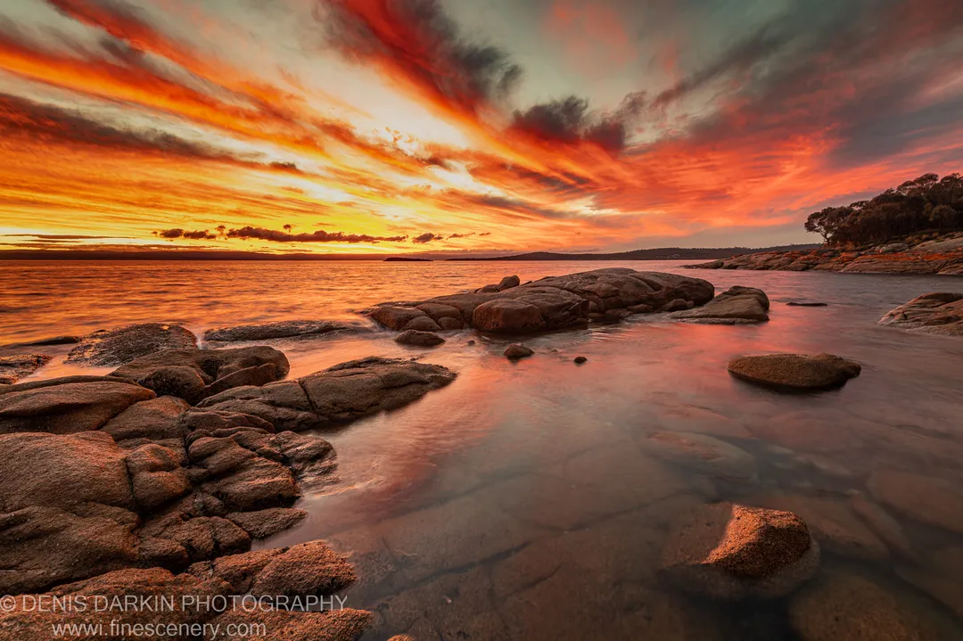 Smooth stones and a rocky coastline at Coles Bay, lit by intense orange light as streaky sunset clouds reflect on the calm water.