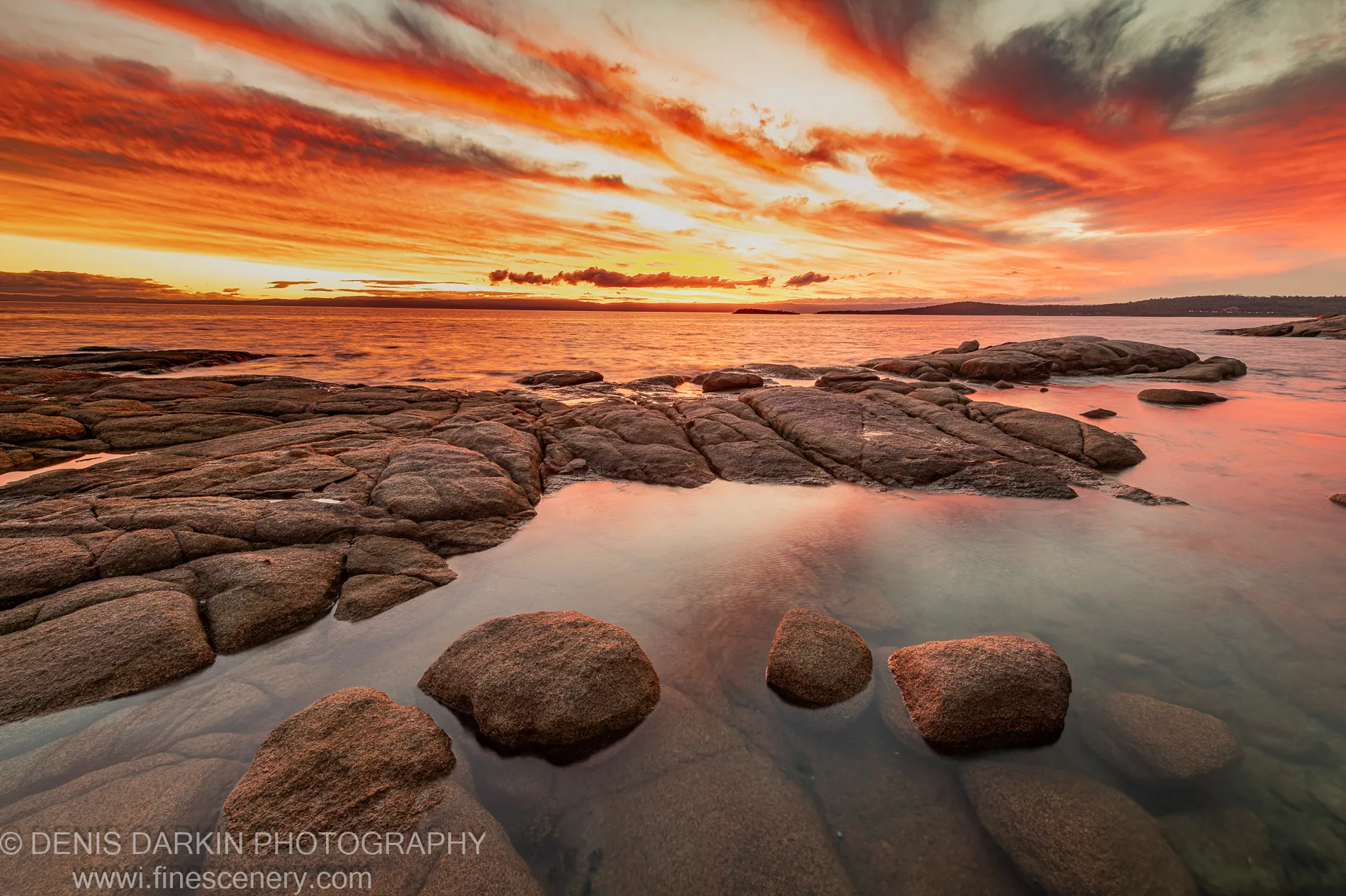 Textured granite boulders rest in shallow water at Coles Bay, reflecting orange and red sunset clouds that stretch across a low horizon.