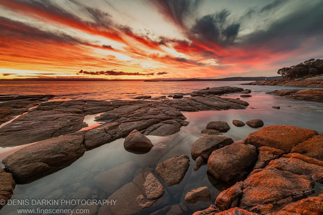 Orange clouds above a granite platform at Coles Bay, with tidal pools reflecting the sunset and storm clouds gathering to the right.