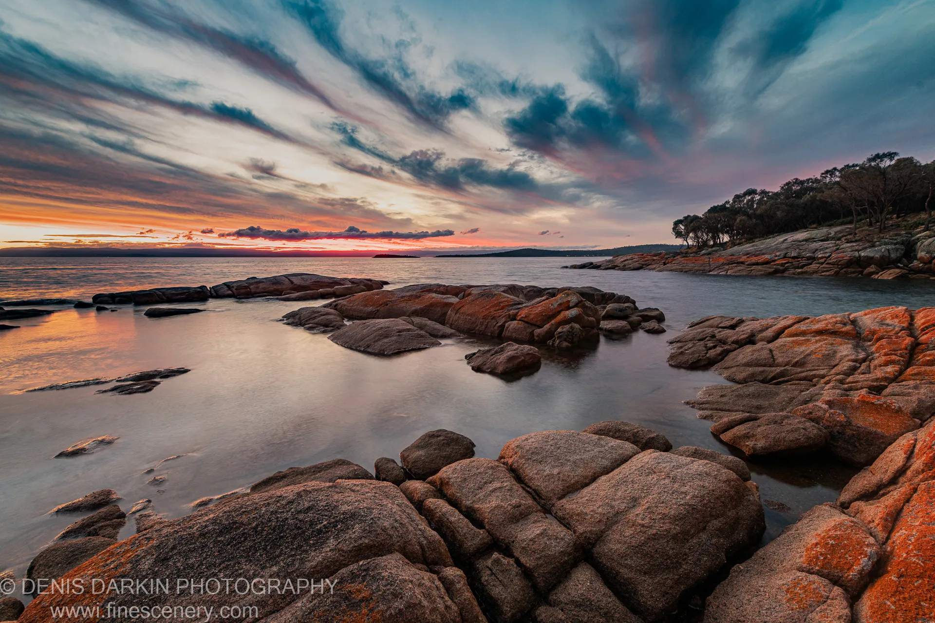 Massive granite boulders rise from long-exposure water at Coles Bay, warm orange sunset light on the rocks and deep blue sky above.