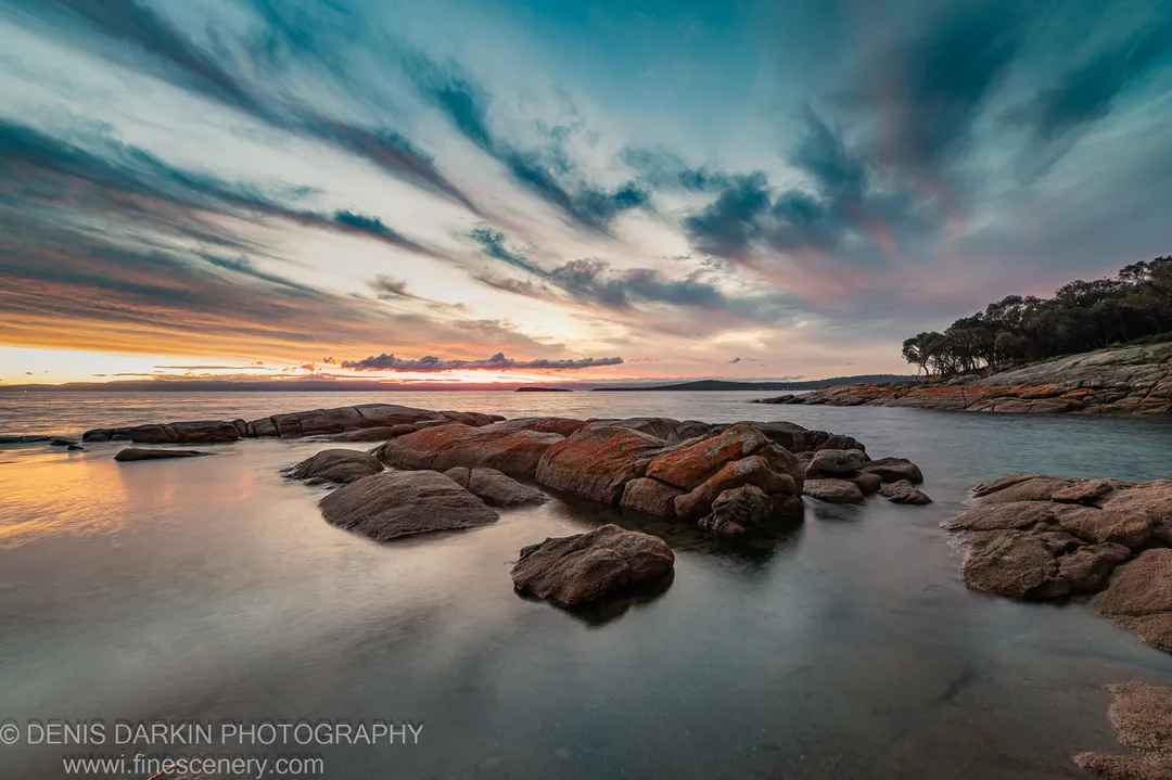 Large reddish granite boulders in long-exposure water at Coles Bay, with intense sunset colors on the left and a treeline on the right.