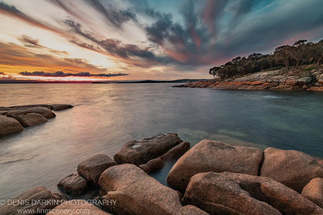 Rounded granite boulders at Coles Bay, with motion-blurred tidal water and a tree-lined headland against orange and blue sunset clouds.
