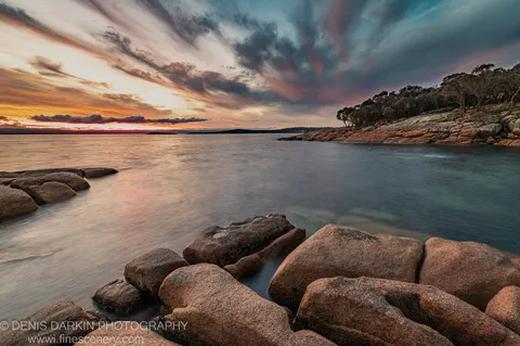 Rounded granite boulders at Coles Bay, with motion-blurred tidal water and a tree-lined headland against orange and blue sunset clouds.