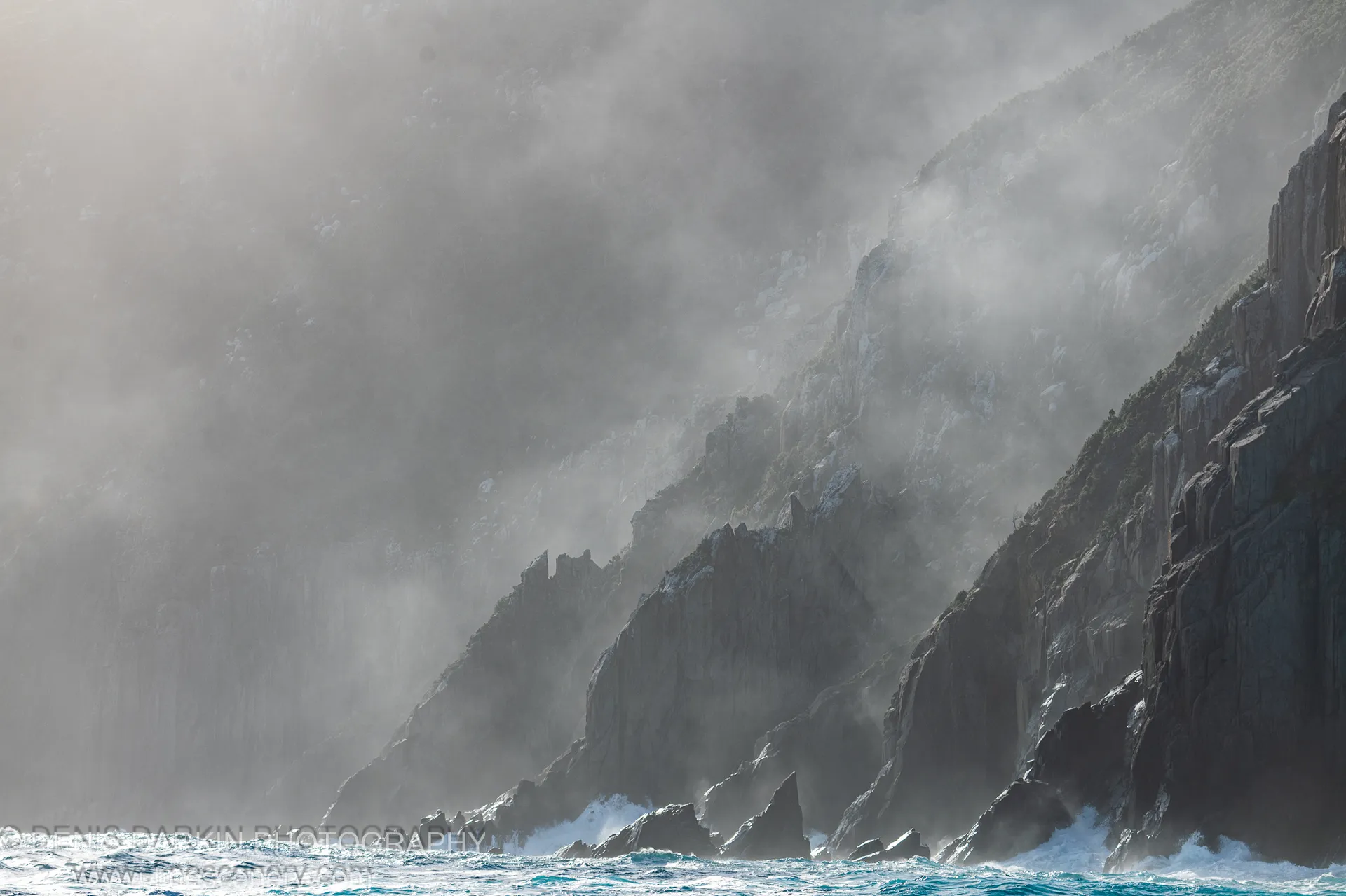Stratified dolerite cliffs at Cape Pillar rise from turbulent water, their vertical faces softened by thick mist and white foam at the base.