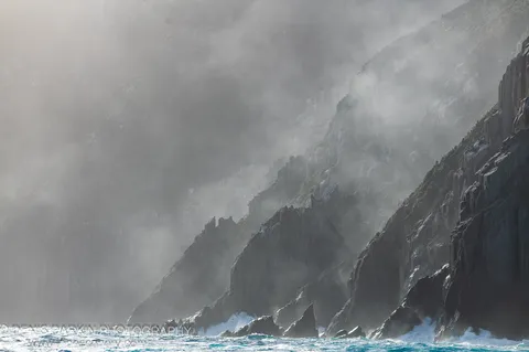 Stratified dolerite cliffs at Cape Pillar rise from turbulent water, their vertical faces softened by thick mist and white foam at the base.