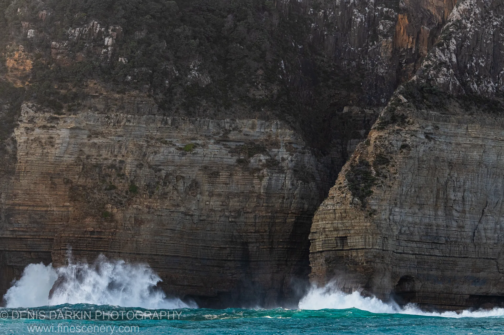 Horizontal cliff layers at Cape Pillar with turquoise waves crashing at the base and soft directional light across the stratified rock surfaces.
