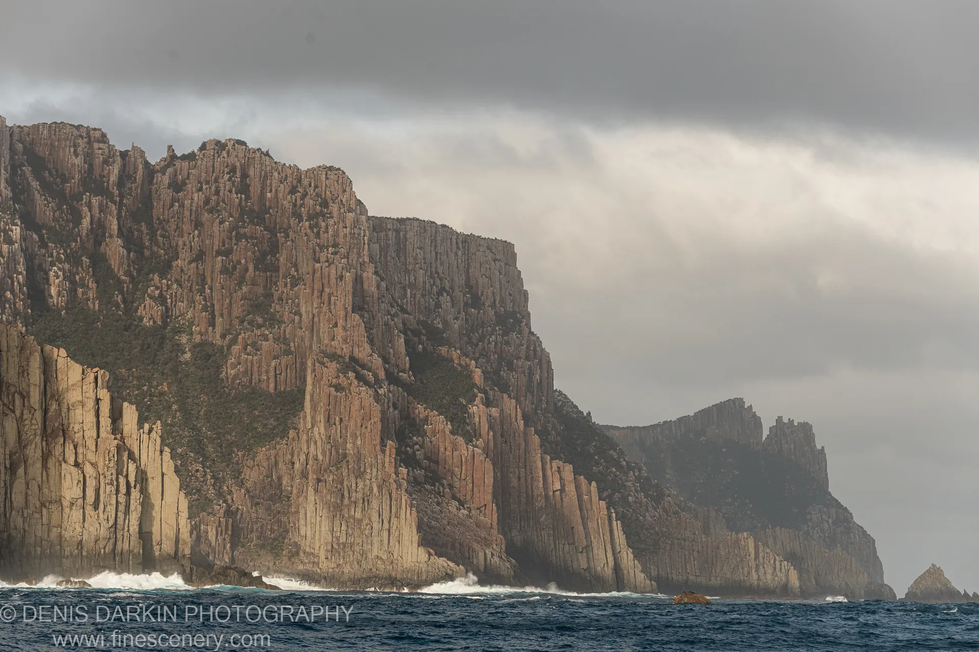 Columnar dolerite cliffs at Cape Pillar rise from a grey sea in diffused light, white foam at the base where waves break.