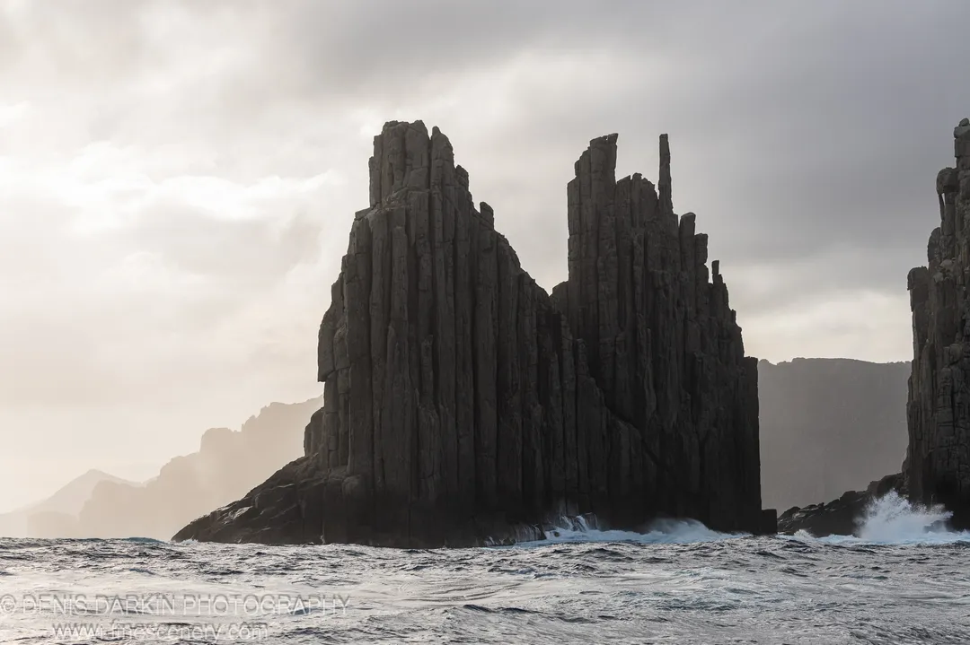 Deep-fissured basalt pillars in soft backlight above crashing white surf at the base of Cape Pillar's sea cliffs, April 2022.