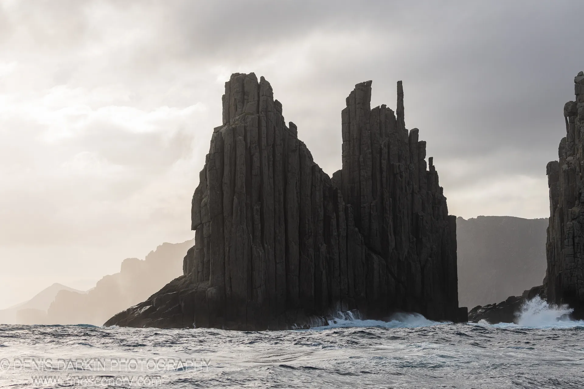 Deep-fissured basalt pillars in soft backlight above crashing white surf at the base of Cape Pillar's sea cliffs, April 2022.