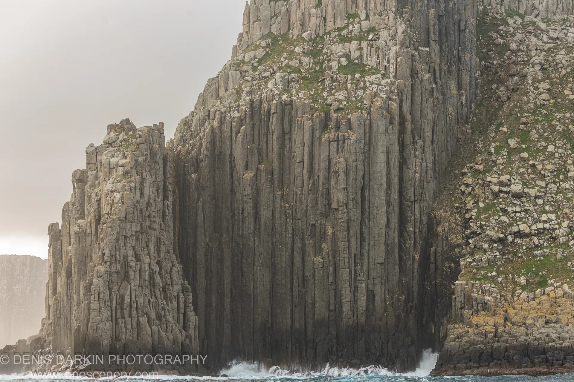 Hexagonal basalt columns rise from breaking surf at Cape Pillar, with patches of green vegetation clinging to the upper ledges, April 2022.
