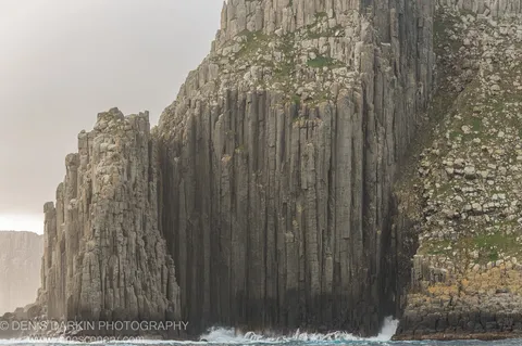 Hexagonal basalt columns rise from breaking surf at Cape Pillar, with patches of green vegetation clinging to the upper ledges, April 2022.