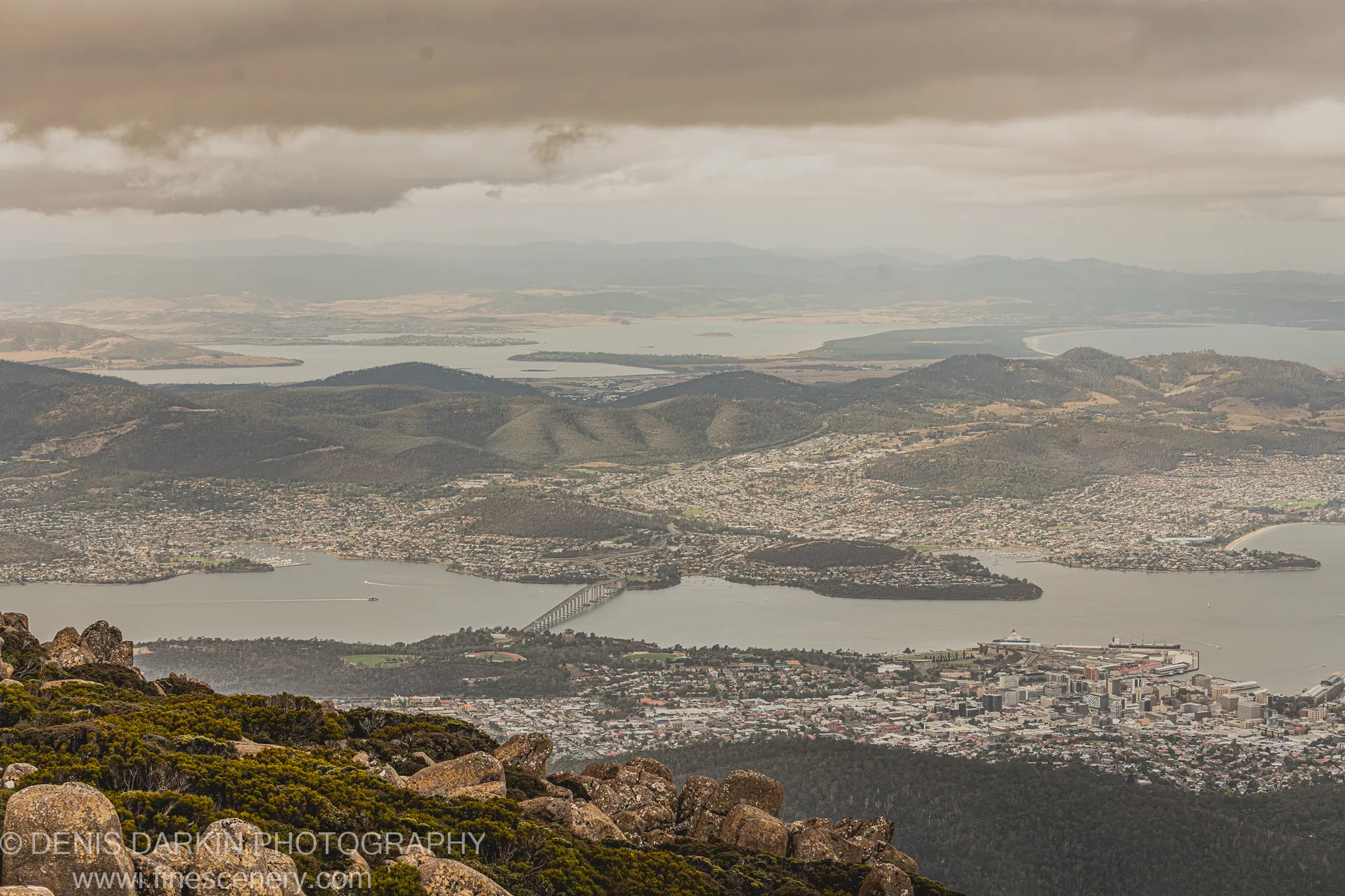 Rough granite boulders and low scrub in the foreground lead toward Hobart on the Derwent Estuary, below heavy storm clouds, April 2022.