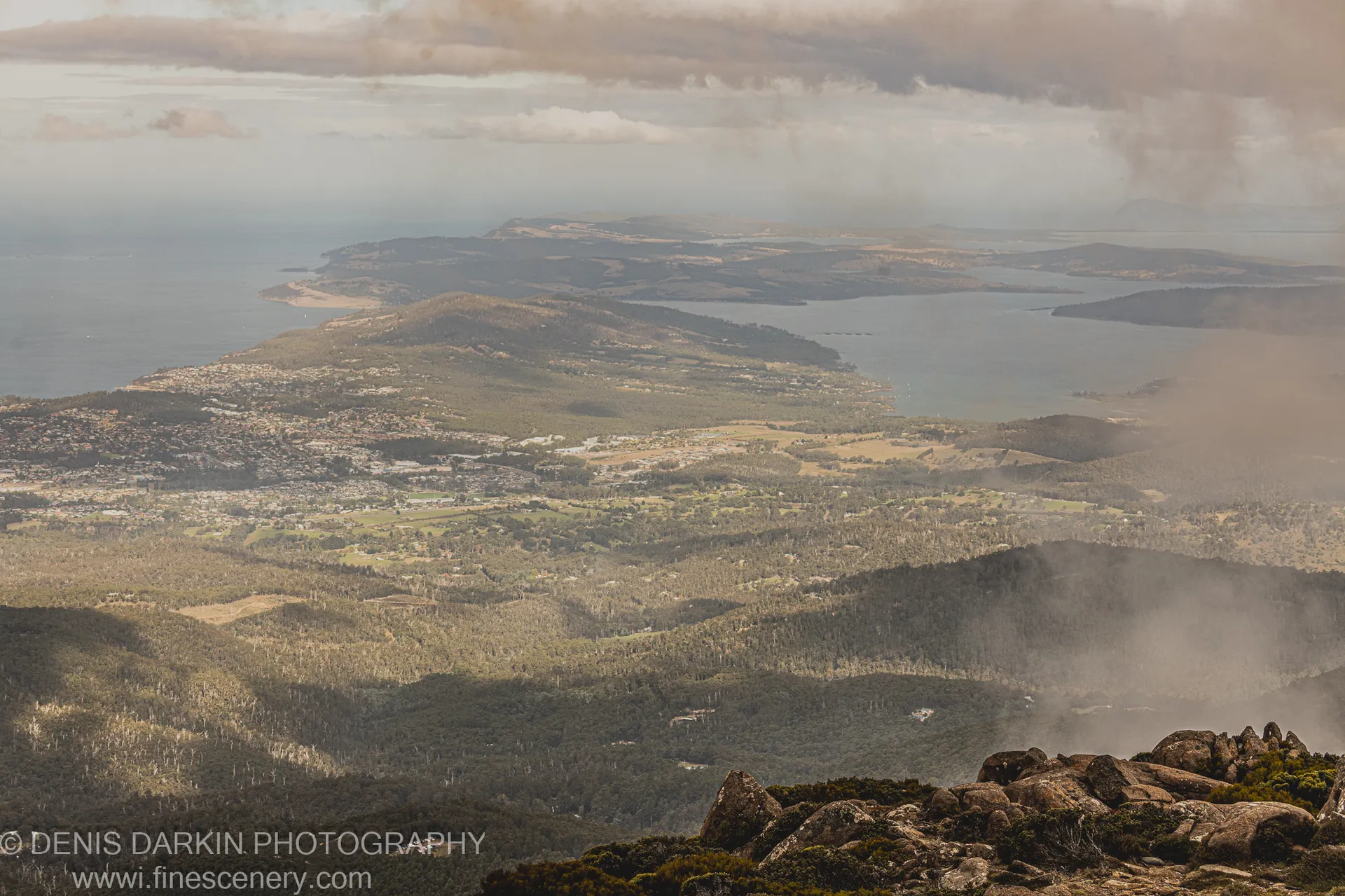 A rugged rocky outcrop in the foreground with Hobart and its surrounding hills extending into coastal haze beyond, April 2022.