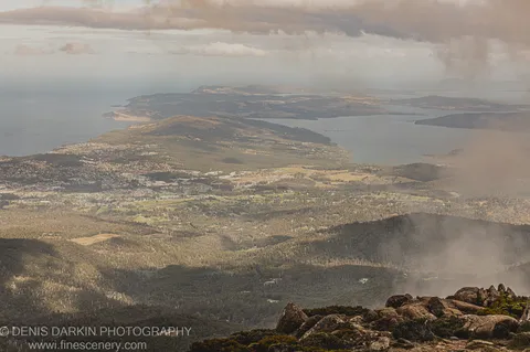 A rugged rocky outcrop in the foreground with Hobart and its surrounding hills extending into coastal haze beyond, April 2022.