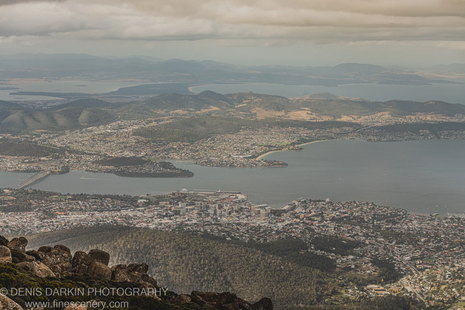 Weathered granite boulders in the foreground anchor a panoramic view of Hobart and the Derwent Estuary below a heavy textured cloud layer, April 2022.