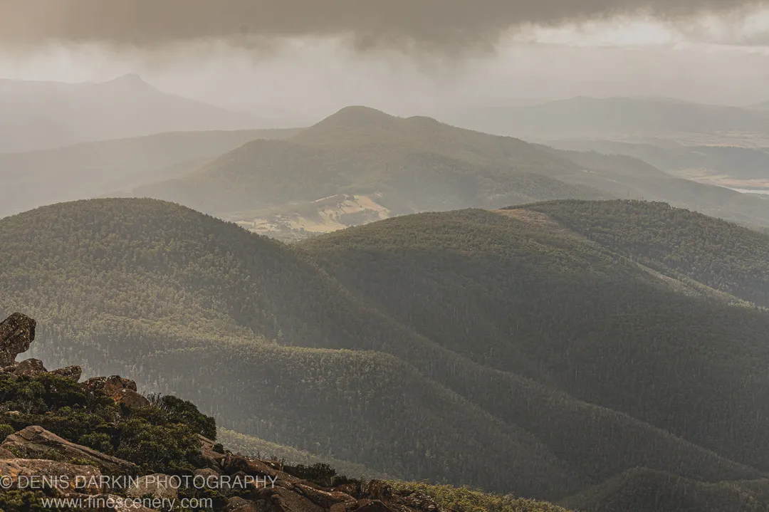 Forested ridges recede into pale autumn haze above a rocky outcrop in the foreground, a wide valley stretching below under overcast sky, Tasmania, April 2022.