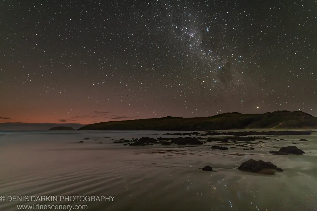 The Milky Way arches over a dark headland at Coffs Harbor, while long exposure smooths the ocean surface around scattered rocks at dawn.