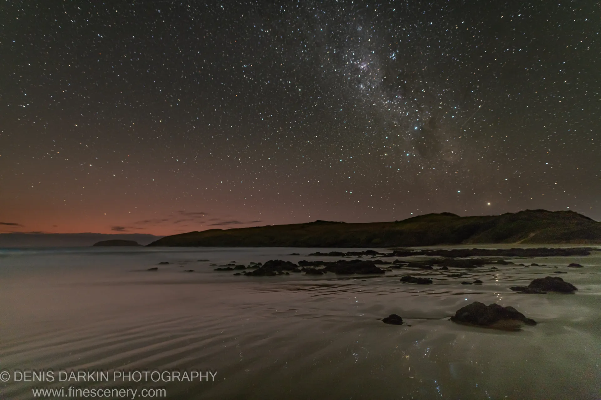 The Milky Way arches over a dark headland at Coffs Harbor, while long exposure smooths the ocean surface around scattered rocks at dawn.
