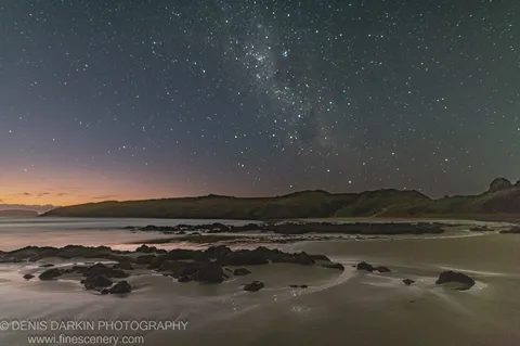 A field of stars fills the sky above silhouetted headlands at Coffs Harbor, with wet sand and jagged rocks in the foreground at dawn.