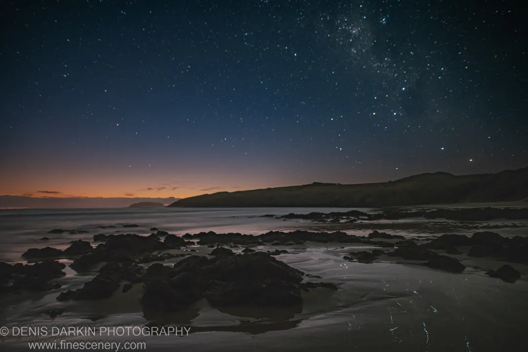 Dark rocks rest on wet sand at Coffs Harbor as a star-filled sky grades into an orange pre-sunrise glow above the still ocean.