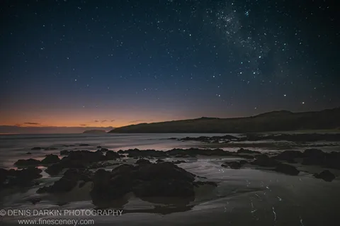 Dark rocks rest on wet sand at Coffs Harbor as a star-filled sky grades into an orange pre-sunrise glow above the still ocean.