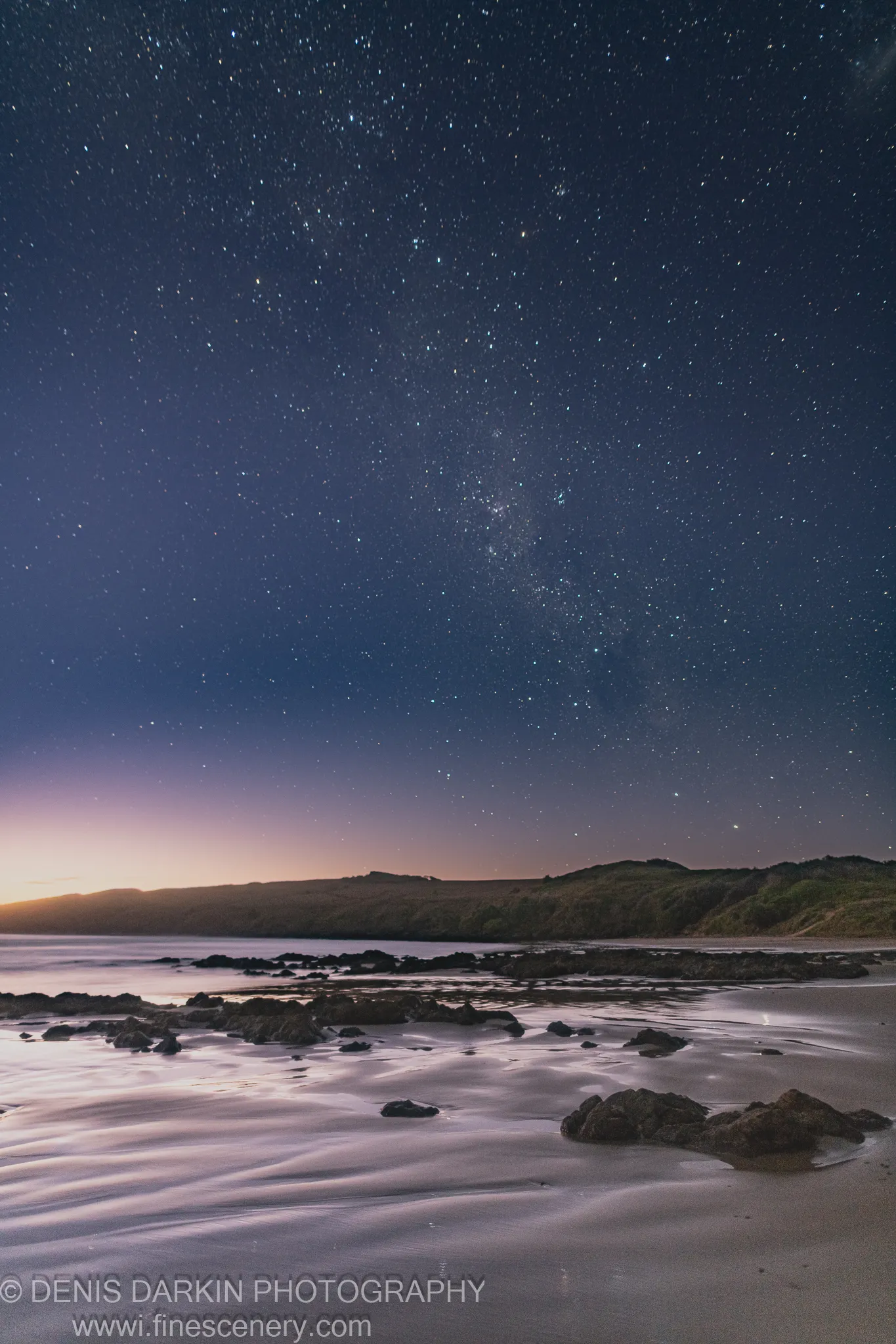 The Milky Way hangs above scattered boulders and rippled sand at Coffs Harbor, with silhouetted dunes in the middle ground at pre-dawn.