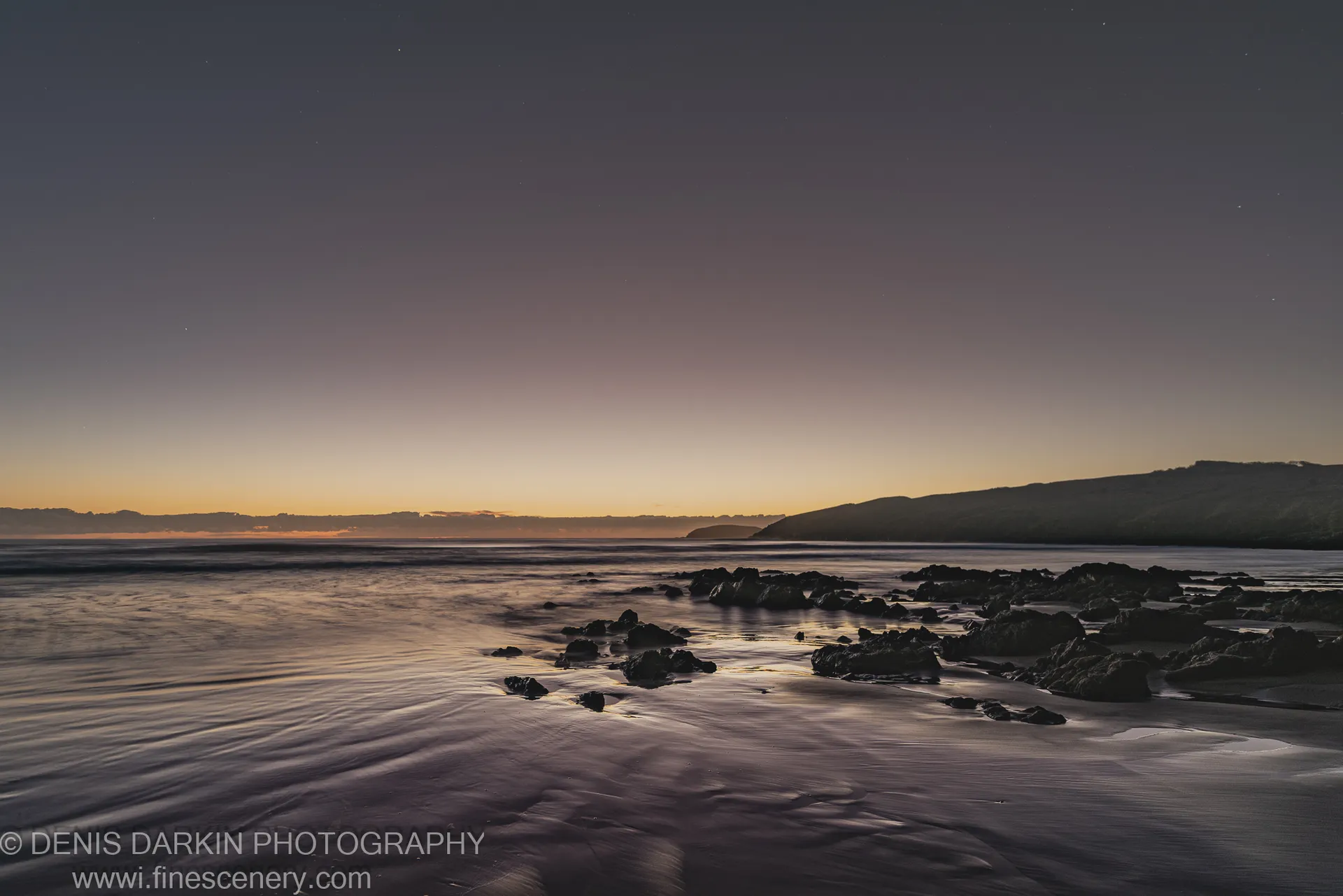 Long-exposure night shot at Coffs Harbor showing wet sand and scattered rocks beneath a sky with faint stars and a warm horizon glow.