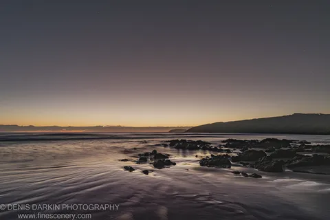 Long-exposure night shot at Coffs Harbor showing wet sand and scattered rocks beneath a sky with faint stars and a warm horizon glow.