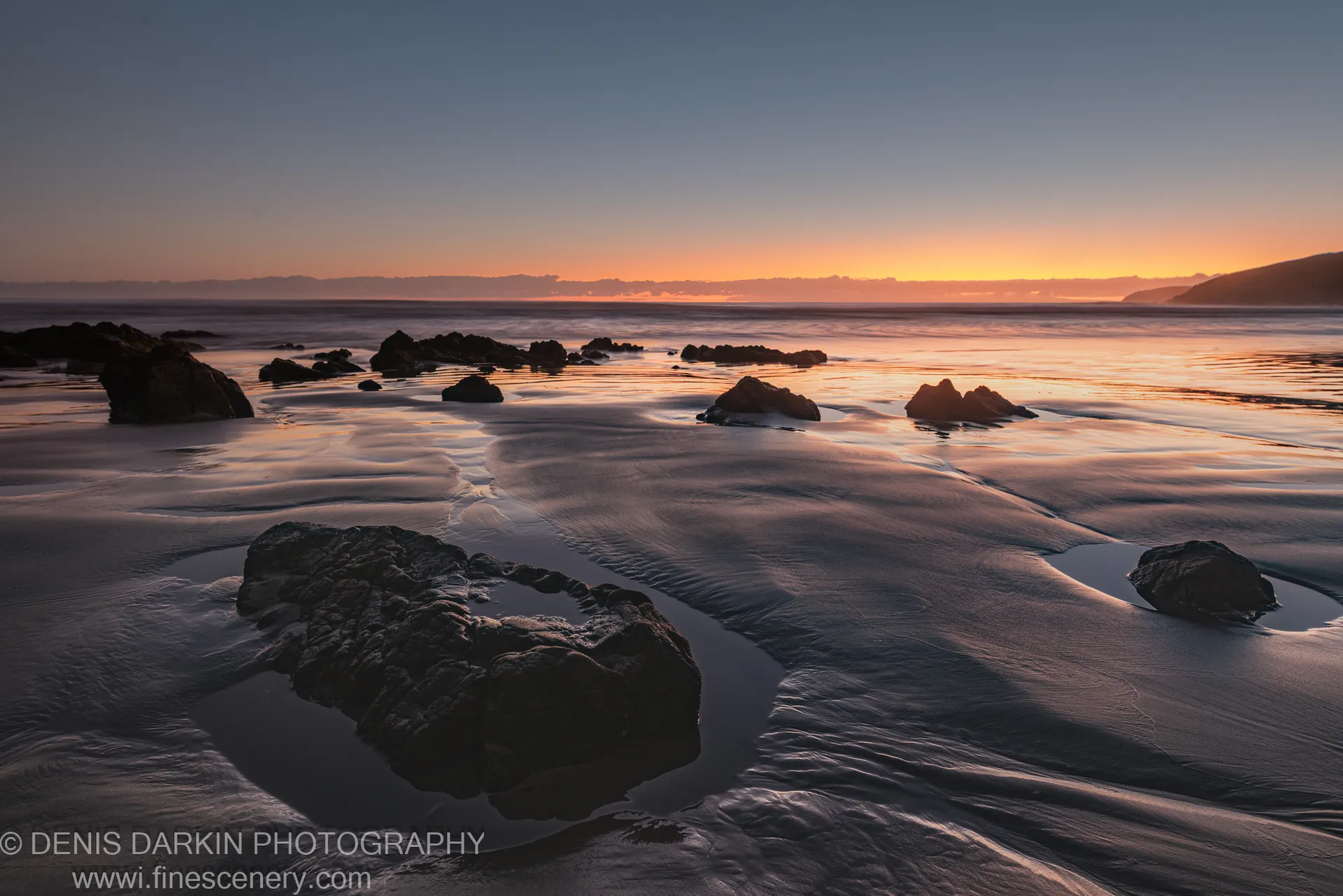 Dark boulders on wet sand at low tide, Coffs Harbor, with long-exposure water and a distant headland visible at night in late autumn.