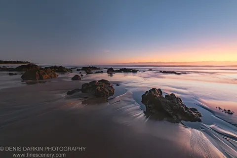 Coastal rocks and wet sand at Coffs Harbor at night, with ripple patterns reflecting an orange glow near the horizon in late autumn.