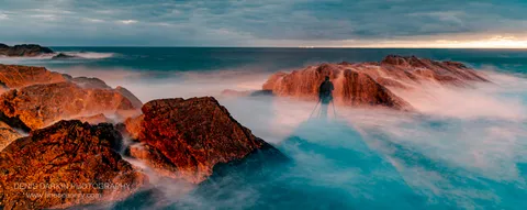 A lone photographer with a tripod stands on coastal rocks at Forster at morning, surrounded by long-exposure surf under a warm morning sky.