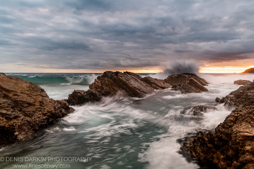 Smooth water swirls around rugged dark rocks on the Forster coast at morning, beneath grey cloud and a warm horizon band.