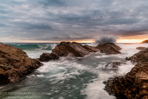 Smooth water swirls around rugged dark rocks on the Forster coast at morning, beneath grey cloud and a warm horizon band.