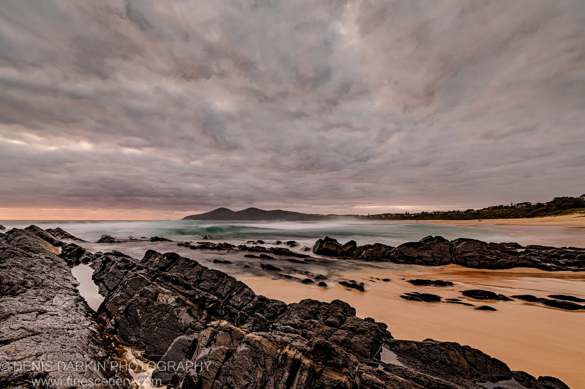 Dark textured rocks overlook a sandy cove at dawn on the Forster coast, warm orange light breaking below heavy grey clouds.