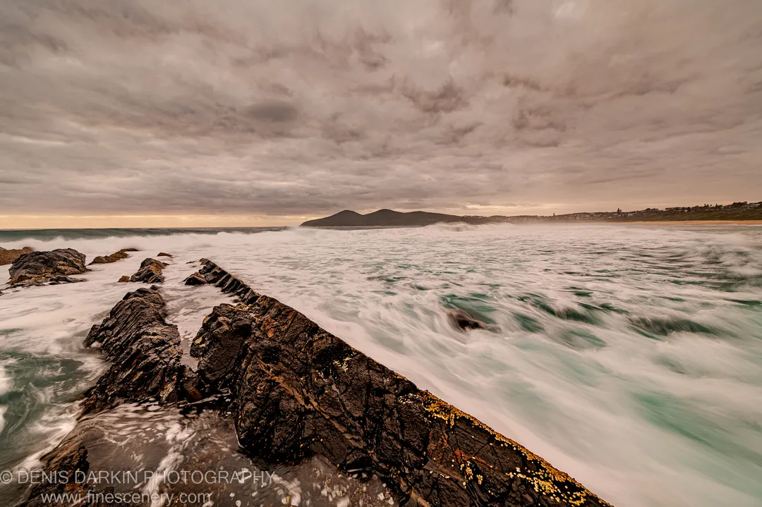 Dark rock formations extend diagonally into long-exposure surf along the Forster coast at dawn, warm horizon light breaking through heavy cloud cover.