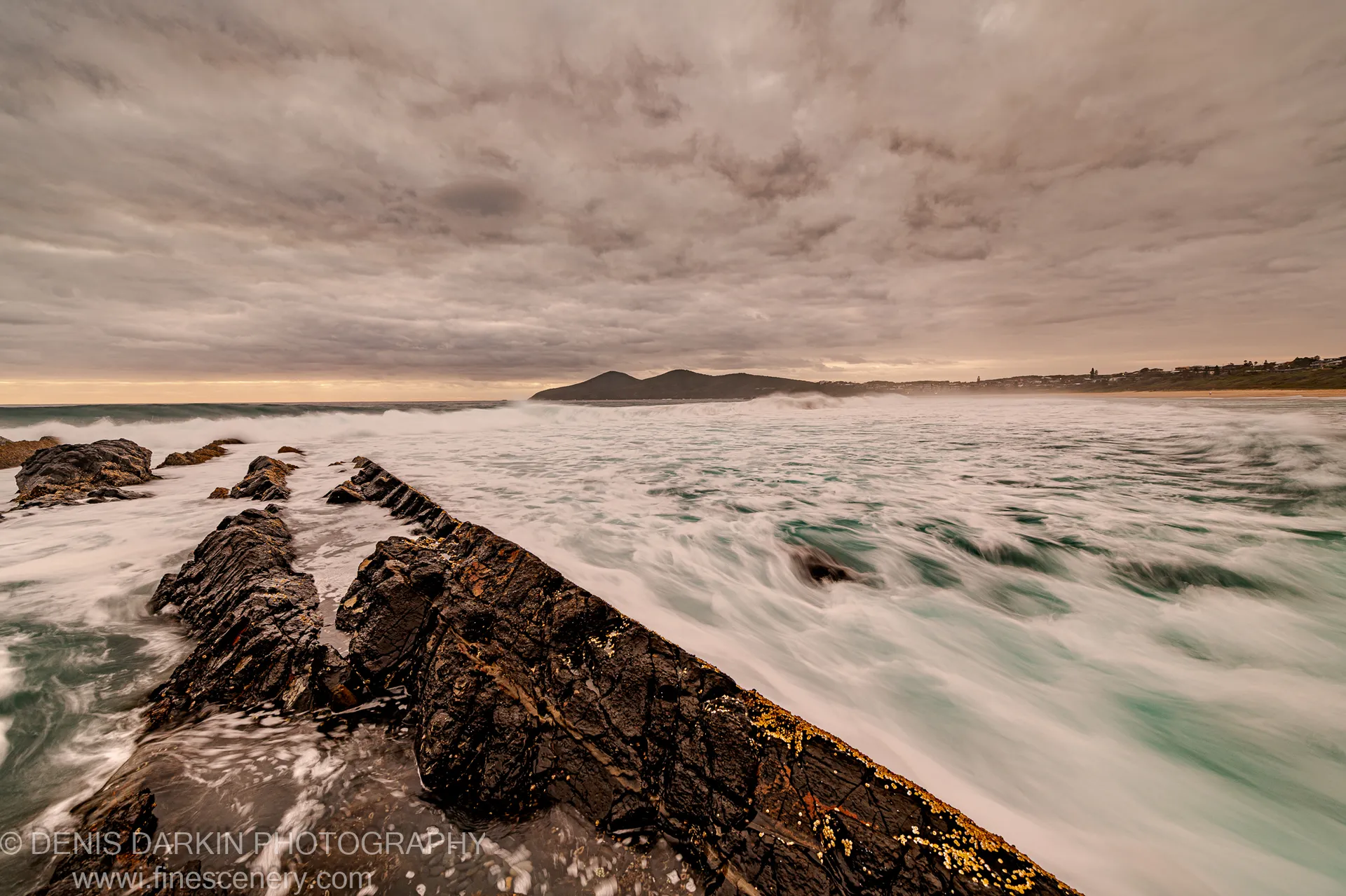 Dark rock formations extend diagonally into long-exposure surf along the Forster coast at dawn, warm horizon light breaking through heavy cloud cover.