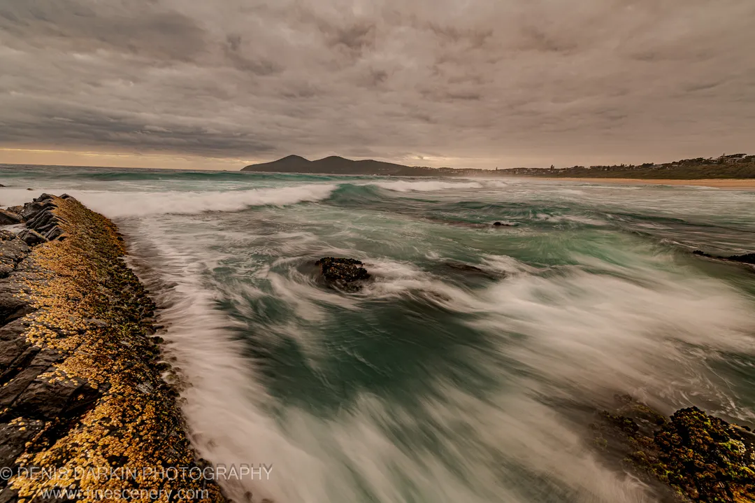 A barnacle-covered jetty at dawn juts into long-exposure surf on the New South Wales coast, with a wide bay visible in the distance.