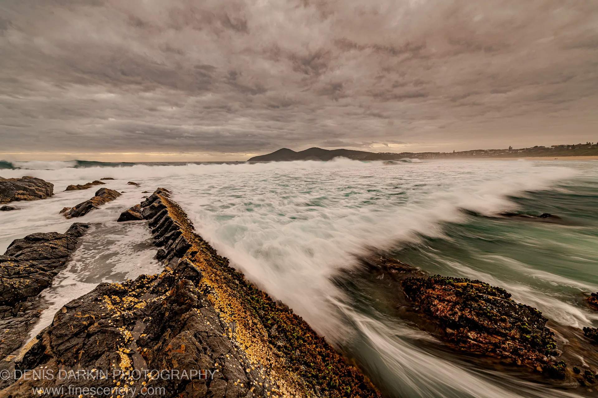 A rocky breakwater with lichen-covered surfaces extends toward a distant headland silhouette under a heavy sky at dawn along the New South Wales coast.
