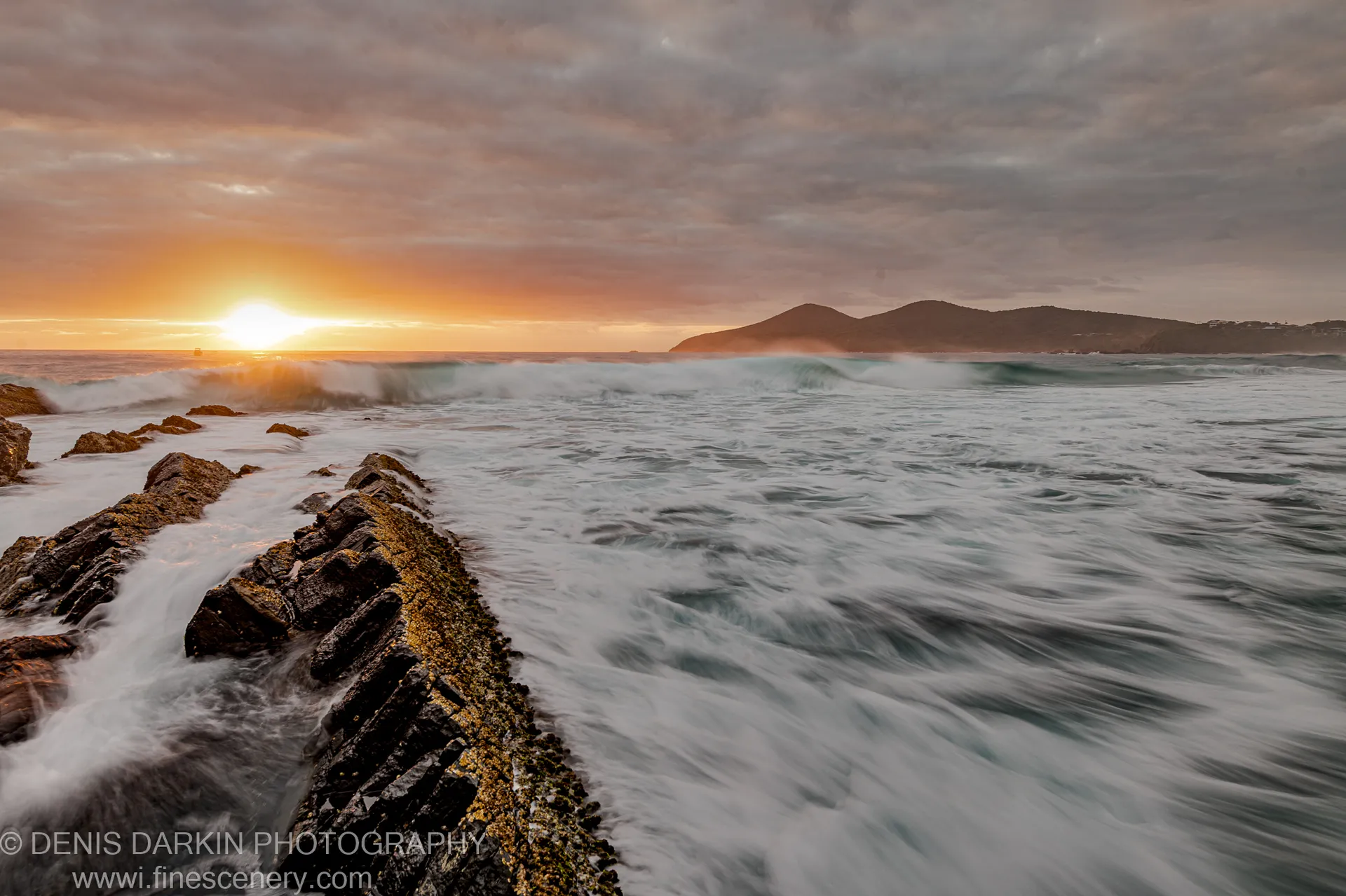 A diagonal stone breakwater juts into long-exposure surf at dawn, warm sunlight catching the wet rock surface under layered morning cloud.