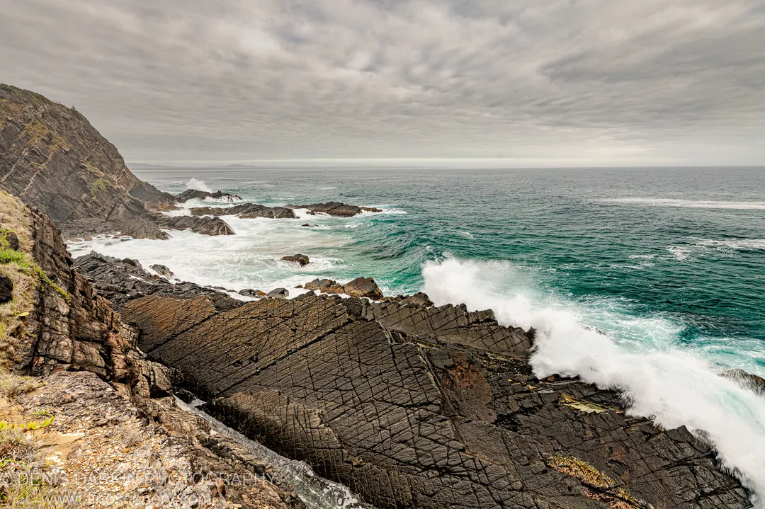Turbulent waves crash against layered rock strata at the Forster headland, white foam rising over the jagged edge under an overcast summer sky.