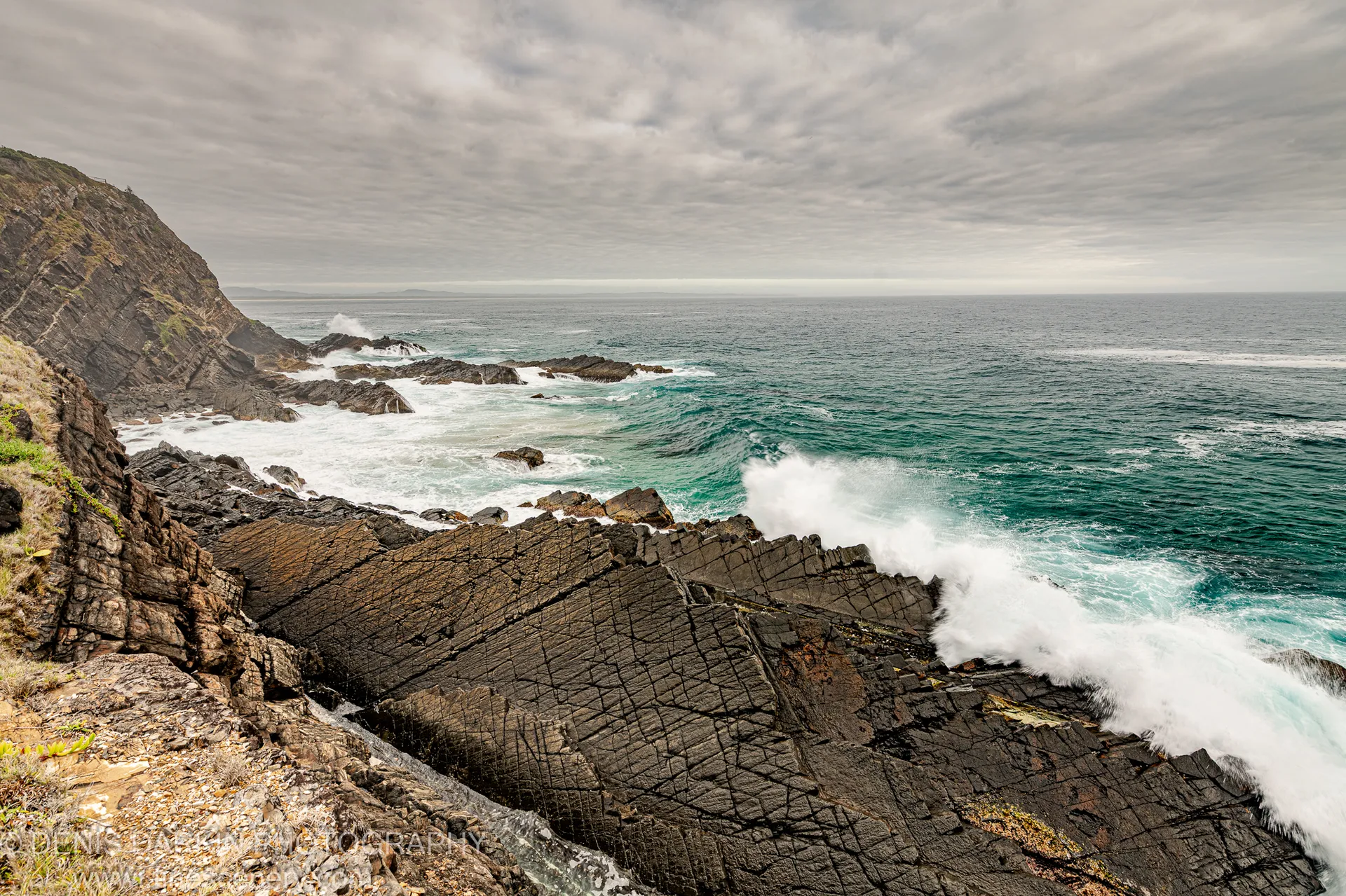 Turbulent waves crash against layered rock strata at the Forster headland, white foam rising over the jagged edge under an overcast summer sky.