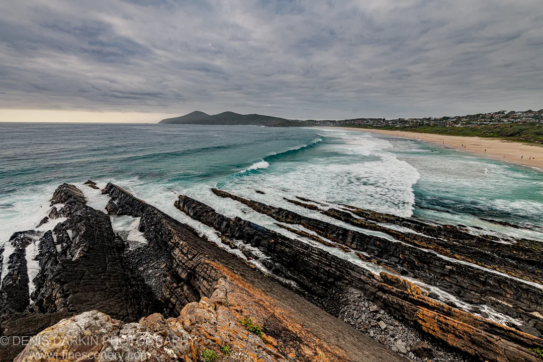 Layered rust-streaked shale and orange lichen in the foreground lead toward breaking waves and a sandy beach arc under a textured sky.