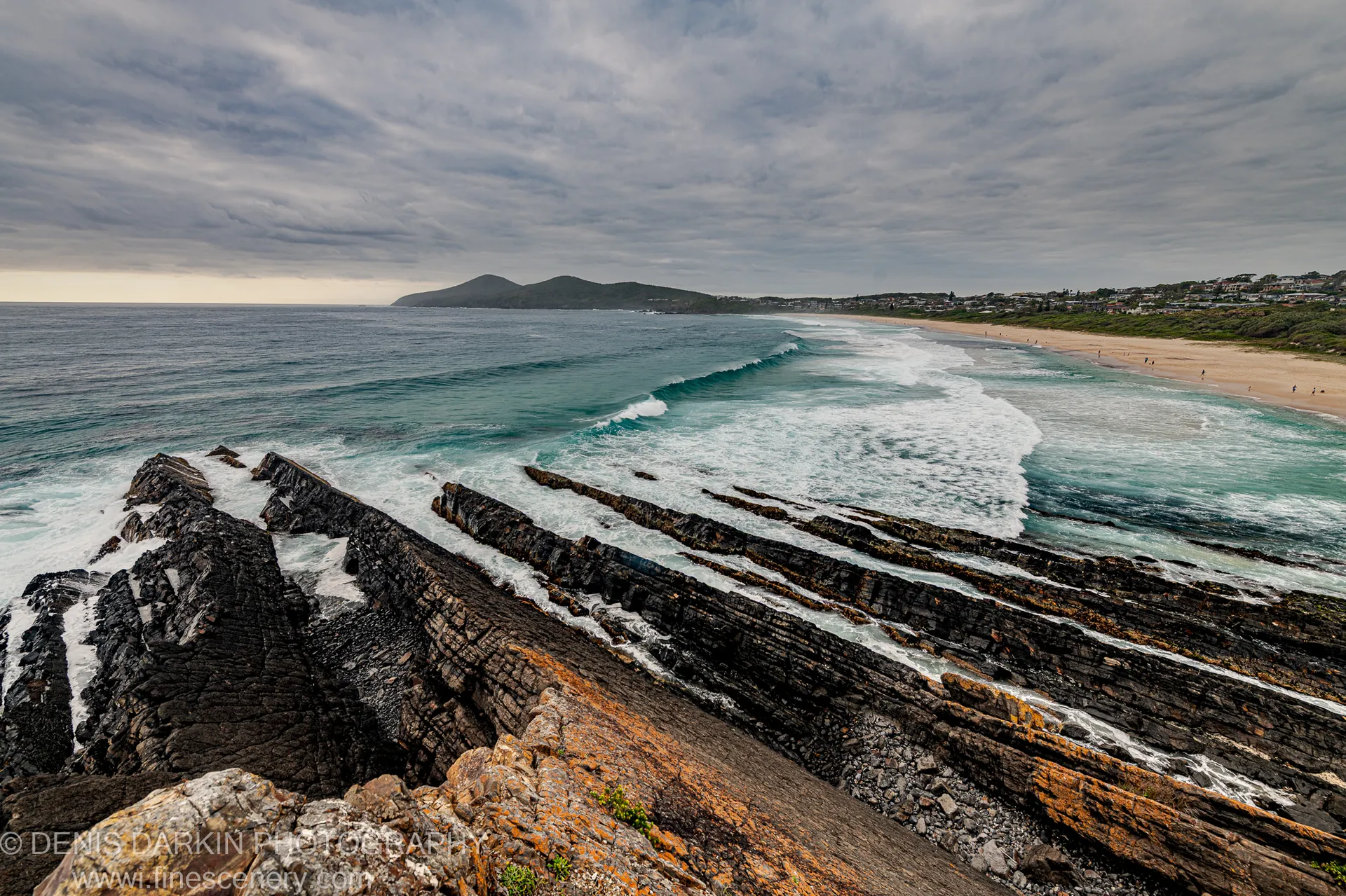 Layered rust-streaked shale and orange lichen in the foreground lead toward breaking waves and a sandy beach arc under a textured sky.